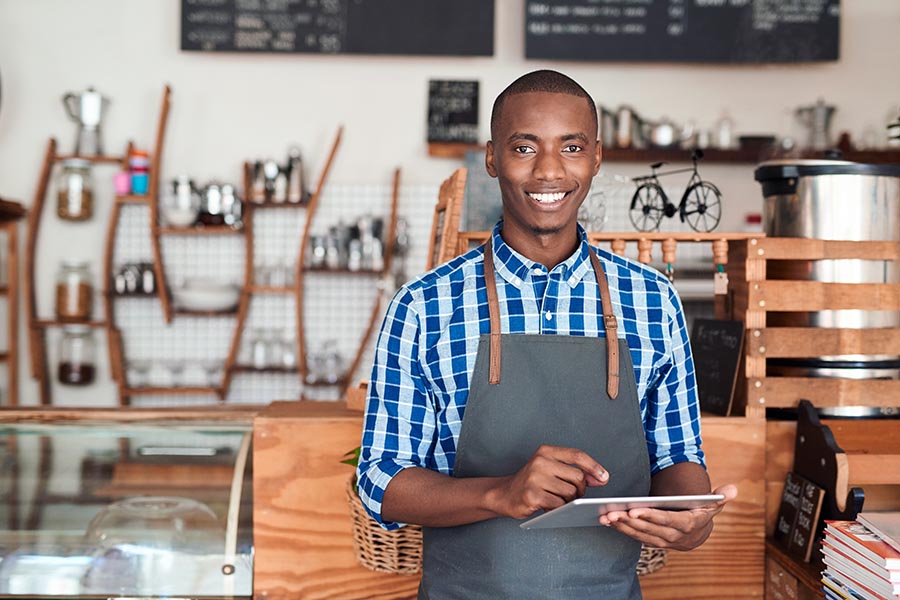 Business Insurance - Business Owner Uses a Tablet While Wearing a Denim Apron, Standing in Front of His Shop Counter