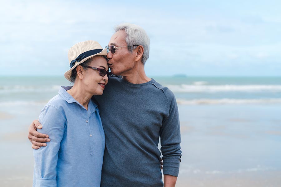 Medicare - Senior Couple Walks on a Beach, Husband Kissing Wife on the Forehead